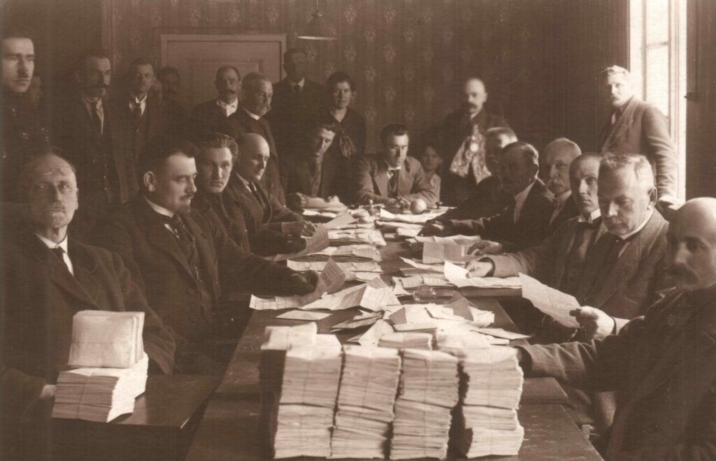 Lai viss būtu likumīgi.  Eduards Radziņš (2.no kreisās) – 1922.gada pašvaldības vēlēšanu biļetenu skaitīšanas komisijā. Aizmugurē stāv slavenais Šana jeb Kārlis Konrāds (ar īpašo medaļu)