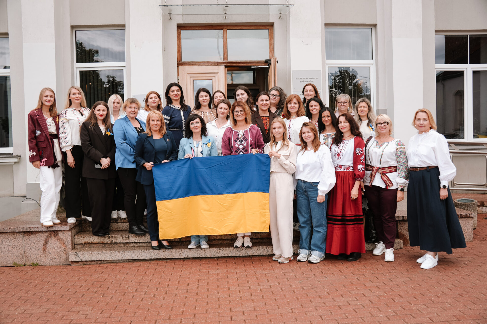 Ukrainas uzņēmēju vizīte Valmierā. Stāv sievietes pie Valmieras novada pašvaldības ar Ukrainas karogu rokās.