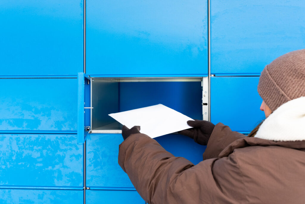 Persona brūnā ziemas jakā un cimdos liek baltu aploksni vai sūtījumu atvērtā zilā pakomāta nodalījumā.