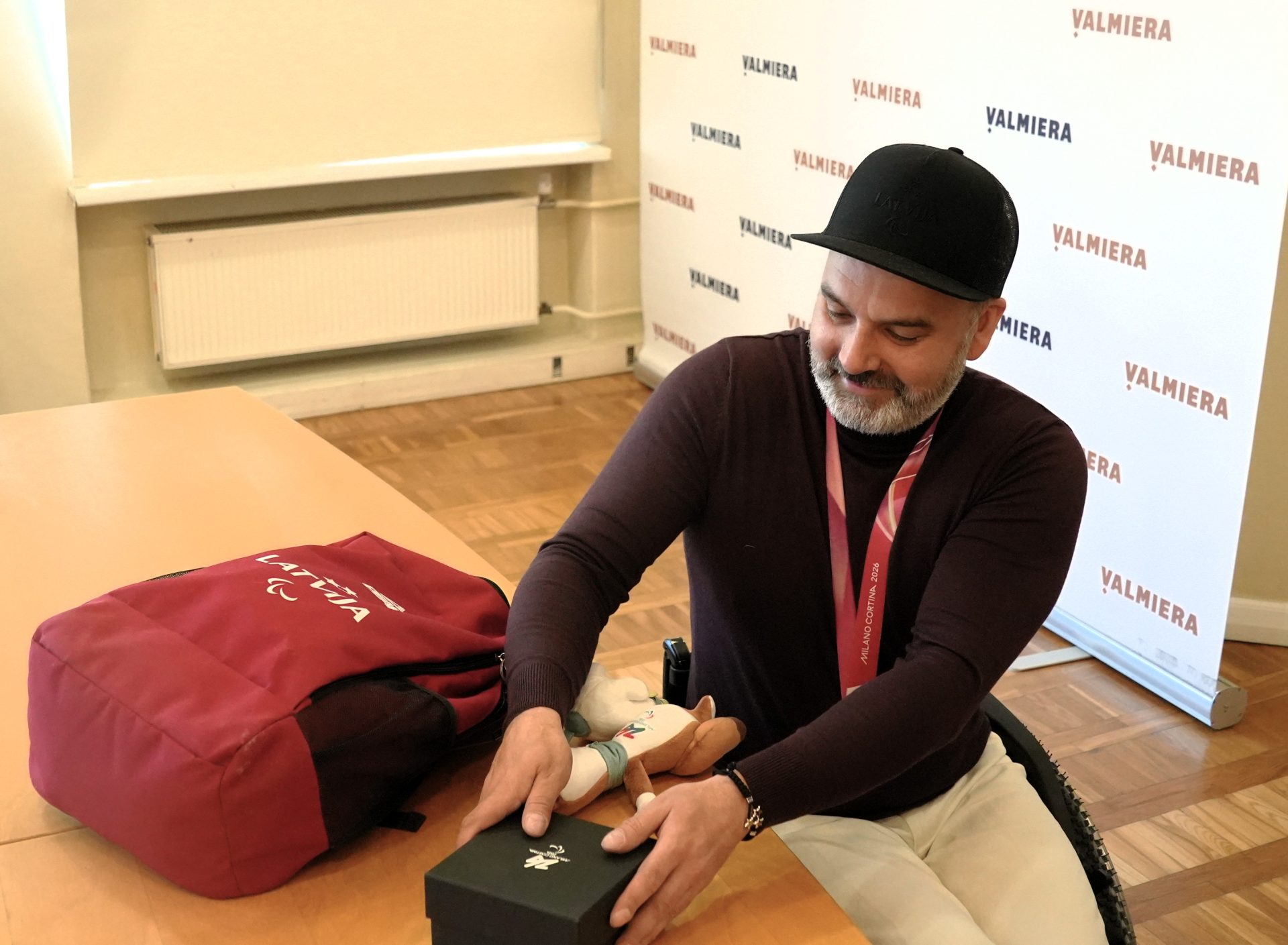 Vīrietis ratiņkrēslā ar medaļu kaklā sēž pie galda un atver melnu dāvanu kastīti; uz galda redzama sarkana soma ar uzrakstu “Latvija”, fonā — Valmieras vizuālā siena.