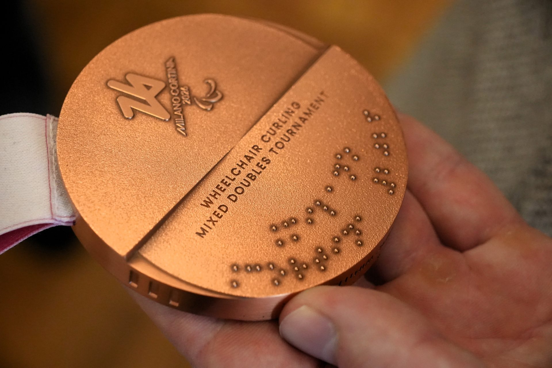Bronzas medaļas tuvplāns plaukstā; uz tās redzams “Milano Cortina 2026” logotips, uzraksts “Wheelchair Curling Mixed Doubles Tournament” un Braila raksts.
