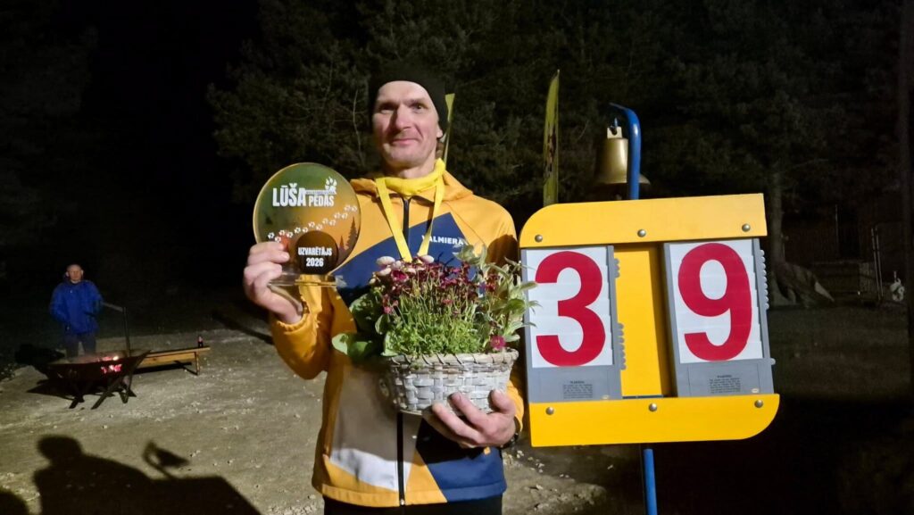 Vīrietis uz tumša meža fona tur rokās balvu “Lūša pēdas”, ziedu grozu un stāv blakus sacensību punktu tablo ar skaitli 39. Fonā redzams ugunskurs un vēl viens cilvēks.