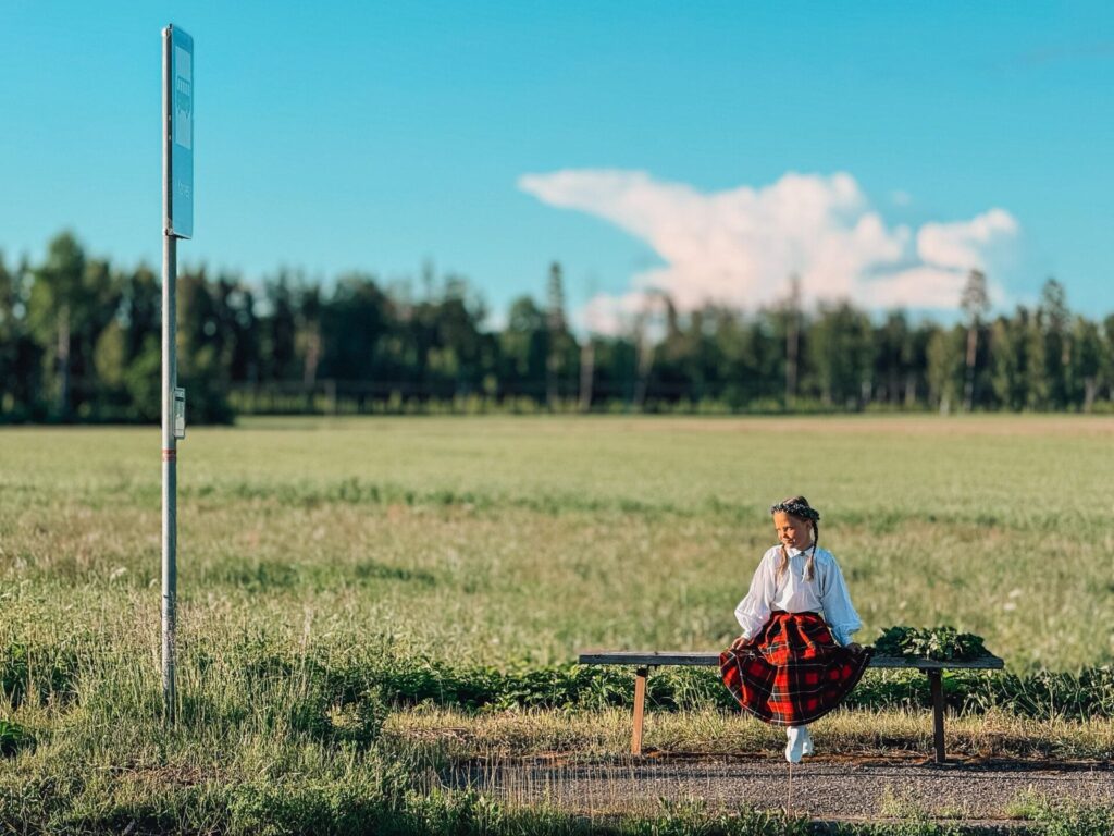 Sabiedriskā transporta pieturā uz soliņa sēž tautas tērptā meitene